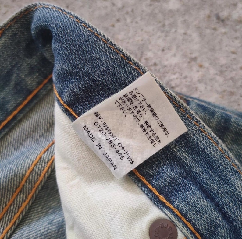 May include: Close-up of a section of blue denim jeans with visible stitching and a white tag. The tag has Japanese text and the words "MADE IN JAPAN". The jeans have a worn appearance.