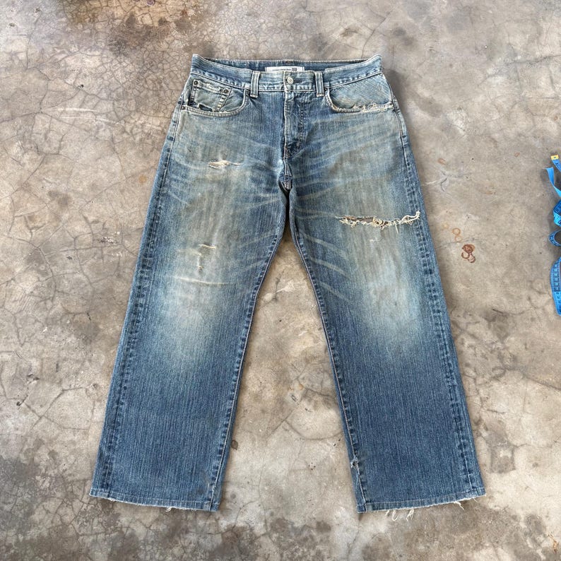 Puede incluir: Vaqueros de mezclilla azul descolorido con un aspecto desgastado. Los vaqueros tienen un dise&ntilde;o cl&aacute;sico de cinco bolsillos, bragueta con botones y un corte relajado. Hay varios rasgu&ntilde;os y roturas en los vaquero, d&aacute;ndoles una apariencia vintage.