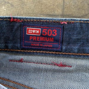 May include: Close-up of a dark blue denim jeans label. The label is navy blue with red and white text that reads "EDWIN 503 PREMIUM MADE IN JAPAN". The jeans have red stitching.