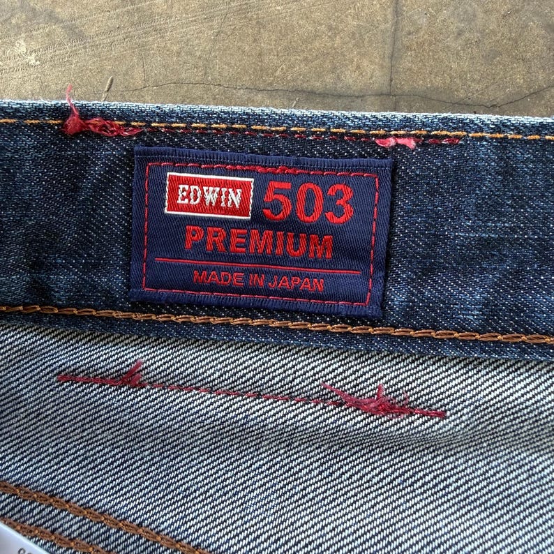 May include: Close-up of a dark blue denim jeans label. The label is navy blue with red and white text that reads "EDWIN 503 PREMIUM MADE IN JAPAN". The jeans have red stitching.