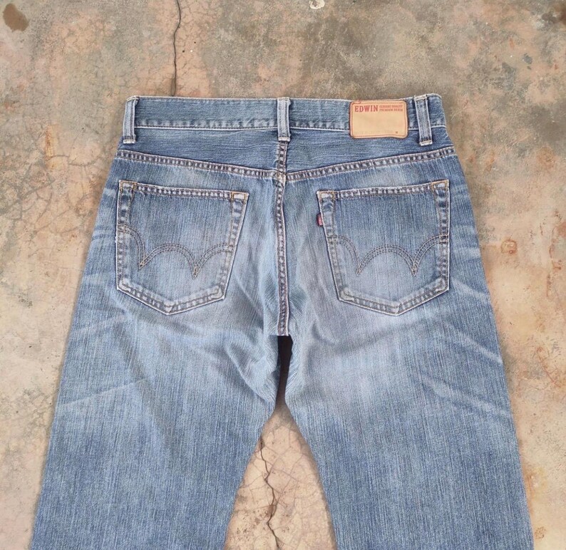 May include: Light blue denim jeans with a classic five-pocket design. The jeans feature a faded wash and visible stitching details. The Edwin brand label is visible on the back waistband. The jeans have a straight leg cut.