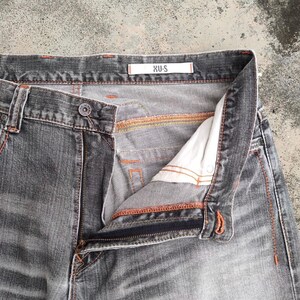 K&ouml;nnte beinhalten: Nahaufnahme einer grauen Jeans mit offenem Rei&szlig;verschluss und Knopfverschluss. Die Jeans hat orangefarbene N&auml;hte und ein wei&szlig;es Etikett mit dem Text "XU-S". Die Jeans ist verblichen und hat ein abgenutztes Aussehen.