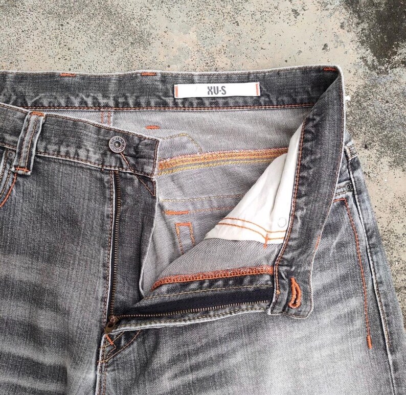K&ouml;nnte beinhalten: Nahaufnahme einer grauen Jeans mit offenem Rei&szlig;verschluss und Knopfverschluss. Die Jeans hat orangefarbene N&auml;hte und ein wei&szlig;es Etikett mit dem Text "XU-S". Die Jeans ist verblichen und hat ein abgenutztes Aussehen.