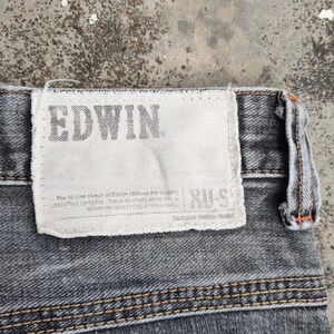 K&ouml;nnte beinhalten: Nahaufnahme eines Abschnitts grauer Jeans. Ein wei&szlig;es rechteckiges Etikett ist auf die Jeans gen&auml;ht, mit dem Markennamen "EDWIN" in Grau aufgedruckt. Die Jeans haben sichtbare N&auml;hte und ein strukturiertes Aussehen.