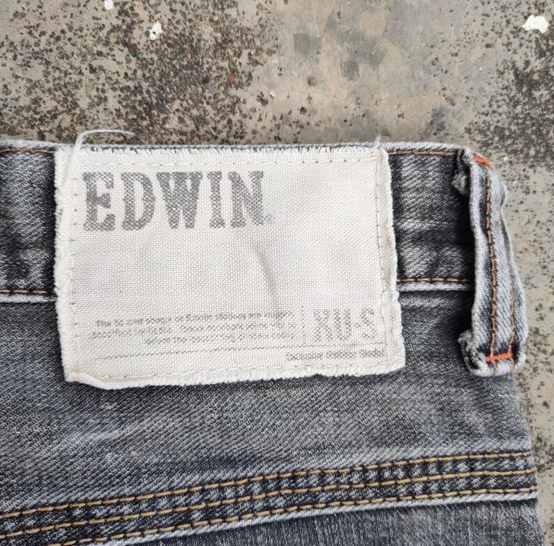 K&ouml;nnte beinhalten: Nahaufnahme eines Abschnitts grauer Jeans. Ein wei&szlig;es rechteckiges Etikett ist auf die Jeans gen&auml;ht, mit dem Markennamen "EDWIN" in Grau aufgedruckt. Die Jeans haben sichtbare N&auml;hte und ein strukturiertes Aussehen.