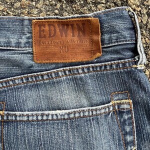 May include: Close-up of blue denim jeans with a brown leather patch. The patch reads "EDWIN EXCLUSIVE VINTAGE" with additional text. The jeans have a classic five-pocket design with visible stitching.