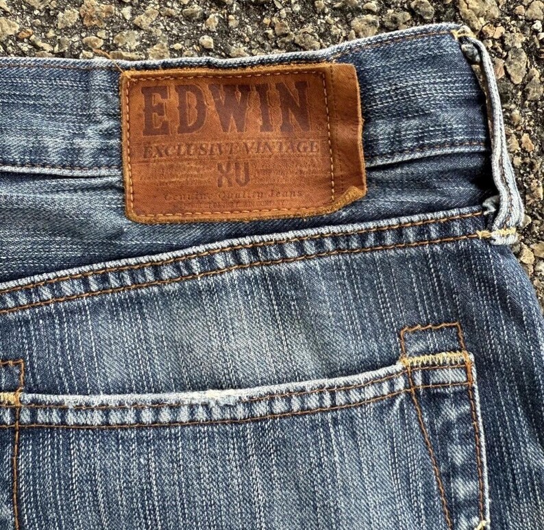 May include: Close-up of blue denim jeans with a brown leather patch. The patch reads "EDWIN EXCLUSIVE VINTAGE" with additional text. The jeans have a classic five-pocket design with visible stitching.