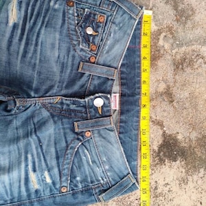 May include: Blue denim jeans with distressed details, featuring classic five-pocket styling and copper-tone hardware. A yellow measuring tape shows a length of approximately 46 cm. The jeans have a button and zipper closure.
