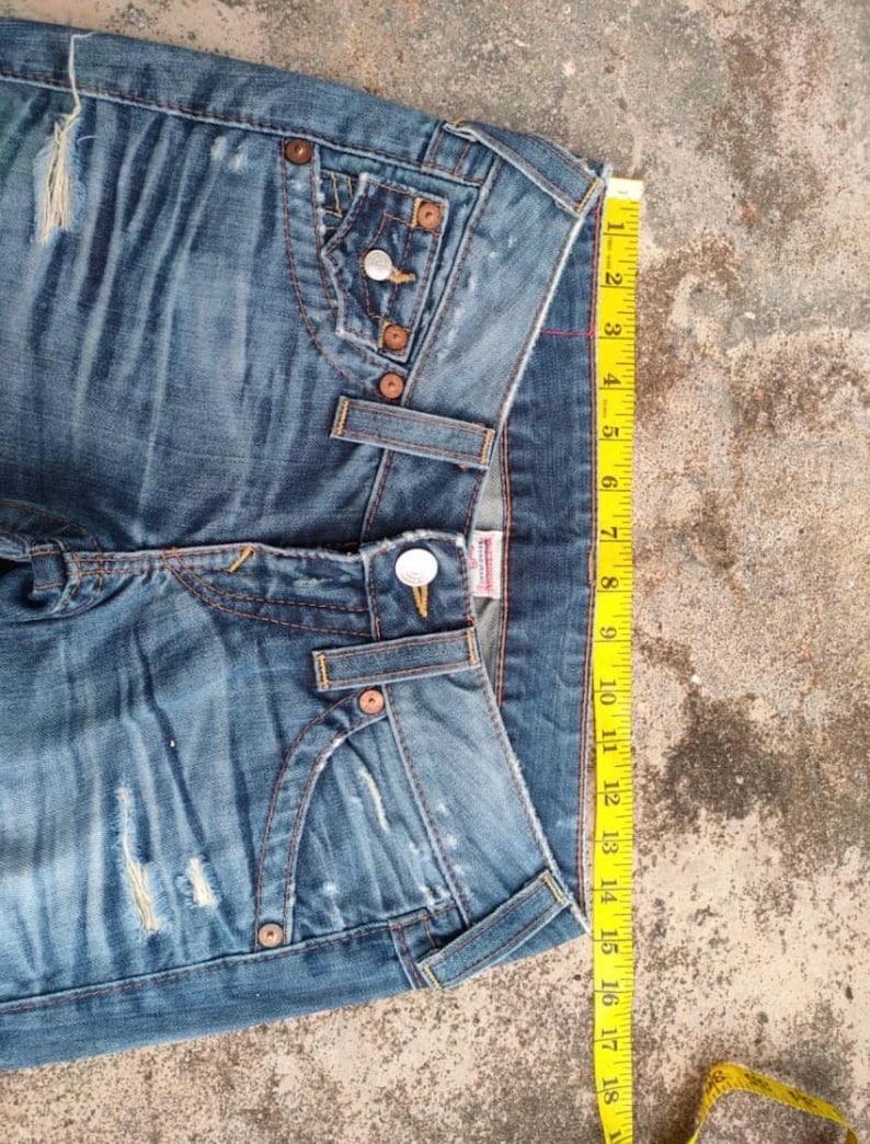 May include: Blue denim jeans with distressed details, featuring classic five-pocket styling and copper-tone hardware. A yellow measuring tape shows a length of approximately 46 cm. The jeans have a button and zipper closure.