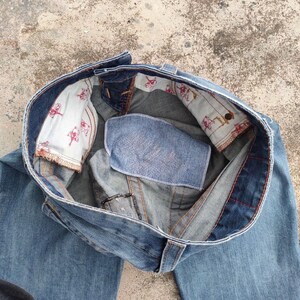 May include: Light blue denim jeans, seen from above, with the waistband open. The interior lining displays a repeating pattern of red figures on a white background. The jeans have a classic five-pocket design and a button closure.