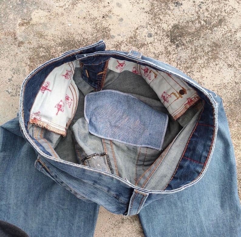 May include: Light blue denim jeans, seen from above, with the waistband open. The interior lining displays a repeating pattern of red figures on a white background. The jeans have a classic five-pocket design and a button closure.