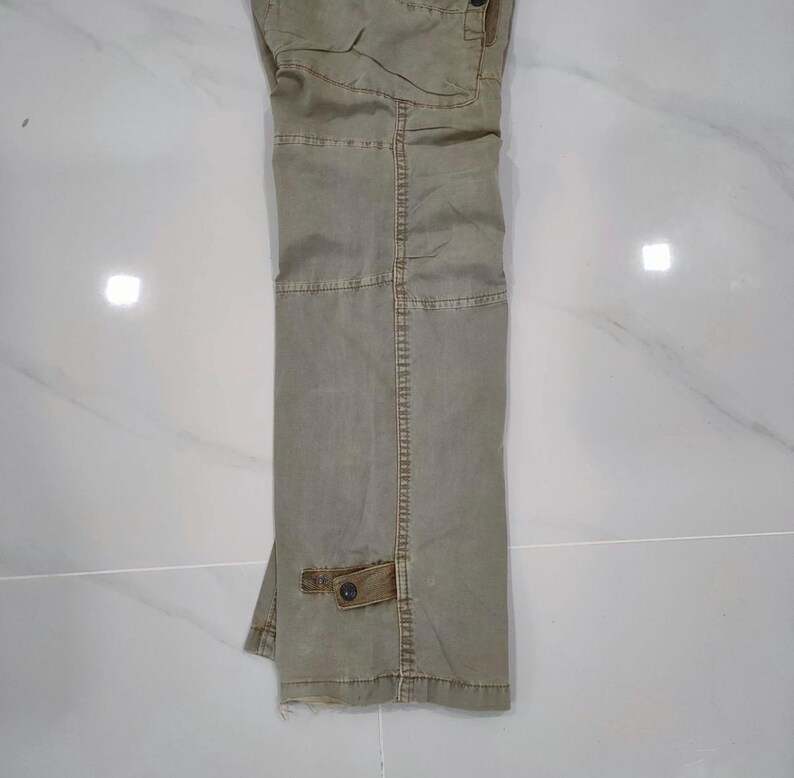 May include: Khaki-colored cargo pants with multiple pockets and a button closure. The pants are made of a durable fabric and have a relaxed fit. The pants are folded and displayed on a white surface.