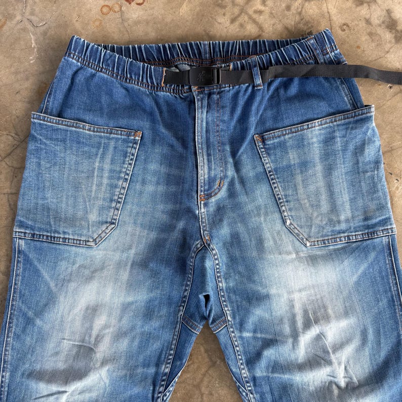May include: Light blue denim pants with large front pockets and a black belt. The pants have an elastic waistband and a faded wash. The pockets are rectangular and stitched with white thread. The belt is made of a black fabric.