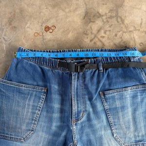 May include: Close-up of blue denim pants with a black belt and large pockets. A blue measuring tape shows a length of approximately 16 inches across the waistband. The jeans have a faded, worn appearance.