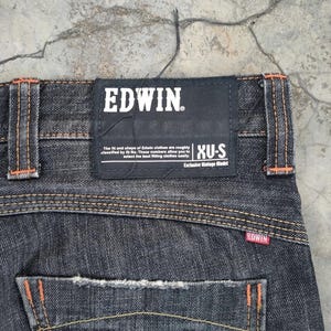 Pu&ograve; includere: Primo piano di jeans di denim scuro con cuciture arancioni e un'etichetta nera Edwin. L'etichetta presenta il nome del marchio in bianco, insieme al testo sulla vestibilit&agrave; e sullo stile. I jeans hanno un classico design a cinque tasche.