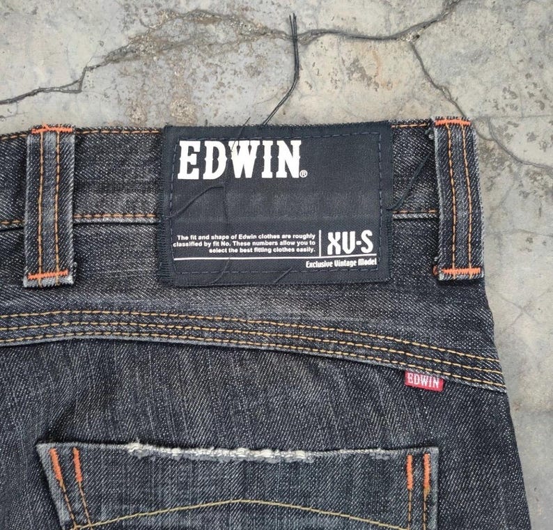 Pu&ograve; includere: Primo piano di jeans di denim scuro con cuciture arancioni e un'etichetta nera Edwin. L'etichetta presenta il nome del marchio in bianco, insieme al testo sulla vestibilit&agrave; e sullo stile. I jeans hanno un classico design a cinque tasche.
