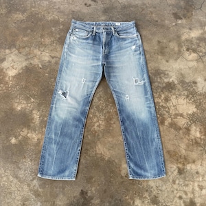 Pu&ograve; includere: Jeans di denim blu chiaro con dettagli strappati, tra cui strappi e sbiaditure. I jeans hanno un classico design a cinque tasche, una chiusura con bottoni e un taglio dritto. I jeans sono appoggiati su una superficie di cemento.