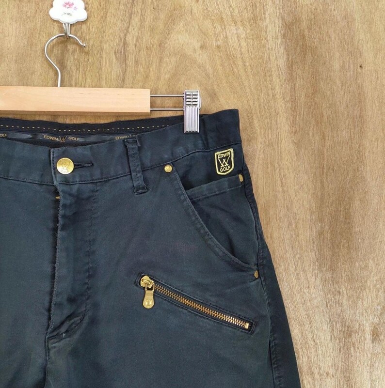 Pu&ograve; includere: Pantaloni da golf blu navy scuro con dettagli dorati, come cerniera e bottoni. I pantaloni hanno una tasca laterale e una piccola toppa con logo dorato. Appesi a una gruccia di legno.