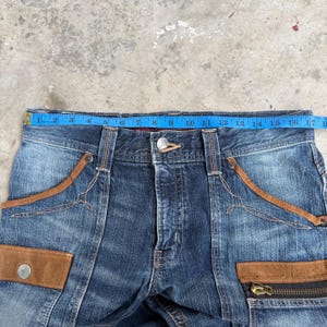 Op de afbeelding: Close-up van een blauwe spijkerbroek met bruine su&egrave;de accenten. De jeans heeft een knoopsluiting, een ritszak en decoratieve stiksels. Een blauwe meetlint toont een lengte van 43 cm.