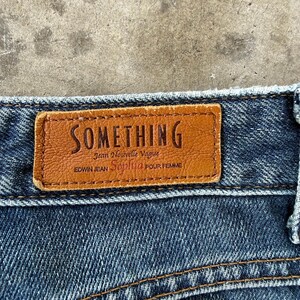 May include: Close-up of a brown leather label stitched onto blue denim jeans. The label reads "SOMETHING" in embossed lettering, with additional text below. The jeans have visible stitching and a classic denim texture.
