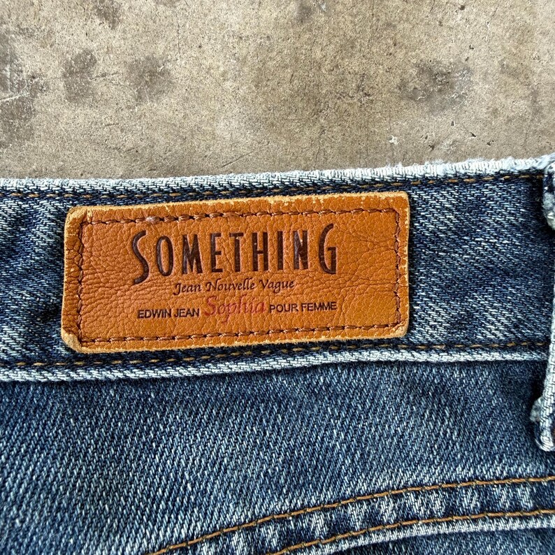 May include: Close-up of a brown leather label stitched onto blue denim jeans. The label reads "SOMETHING" in embossed lettering, with additional text below. The jeans have visible stitching and a classic denim texture.