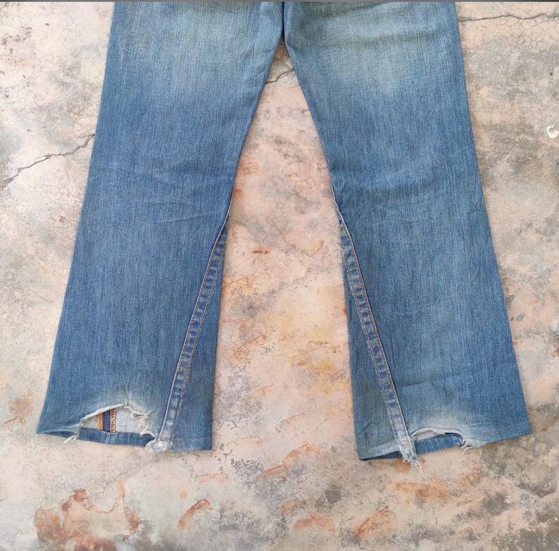 May include: Light blue denim jeans with a flared leg design. The jeans feature a split seam detail on the outer leg and distressed detailing at the hem. The jeans are faded and show signs of wear.