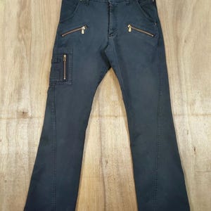 Pu&ograve; includere: Pantaloni blu navy scuro con multiple tasche con cerniera dorate e dettagli. I pantaloni hanno un classico design a cinque tasche con una tasca cargo sulla gamba sinistra. I pantaloni sono appesi a una gruccia di legno su uno sfondo di legno chiaro.