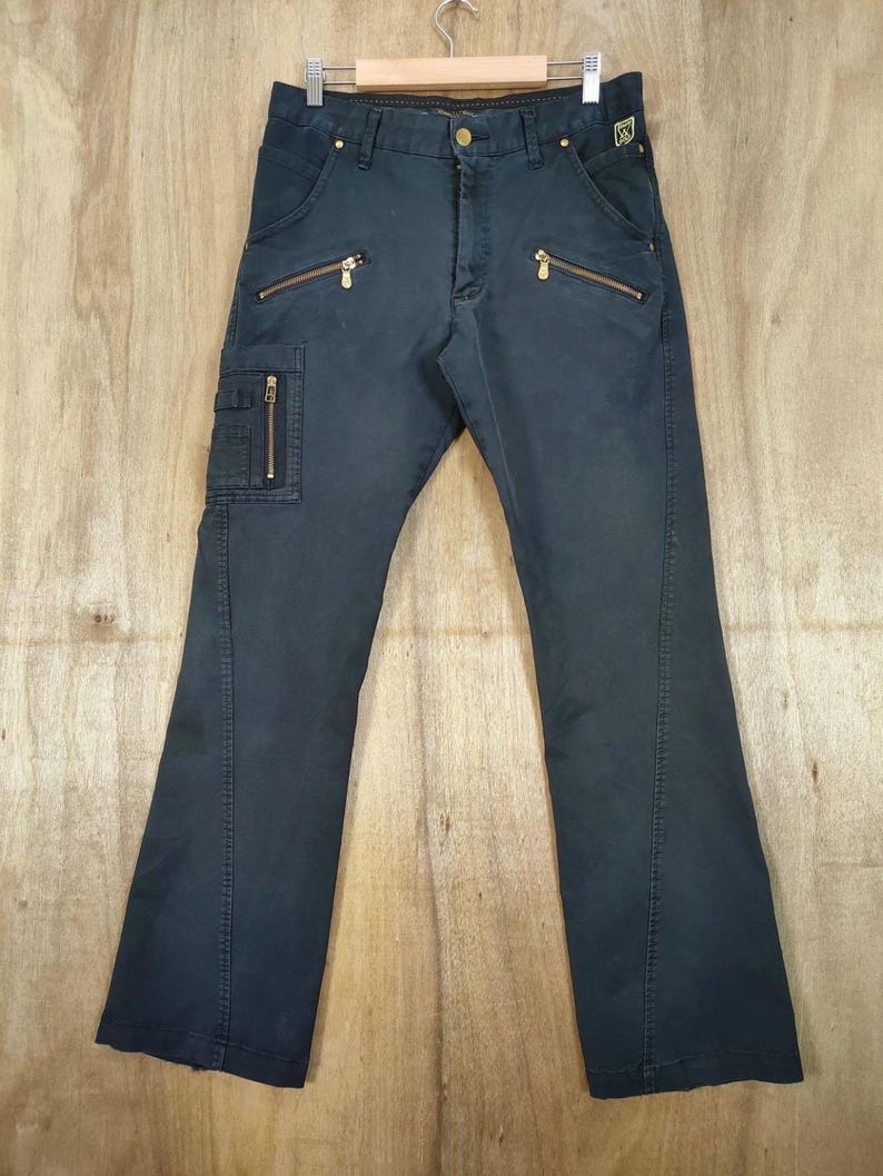 Pu&ograve; includere: Pantaloni blu navy scuro con multiple tasche con cerniera dorate e dettagli. I pantaloni hanno un classico design a cinque tasche con una tasca cargo sulla gamba sinistra. I pantaloni sono appesi a una gruccia di legno su uno sfondo di legno chiaro.