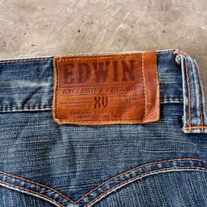 Pu&ograve; includere: Primo piano di jeans di denim blu con cuciture arancioni. Una toppa in pelle marrone con il marchio "EDWIN" e la scritta "EXCLUSIVE VINTAGE" &egrave; cucita sul retro dei jeans. I jeans hanno un classico design a cinque tasche.