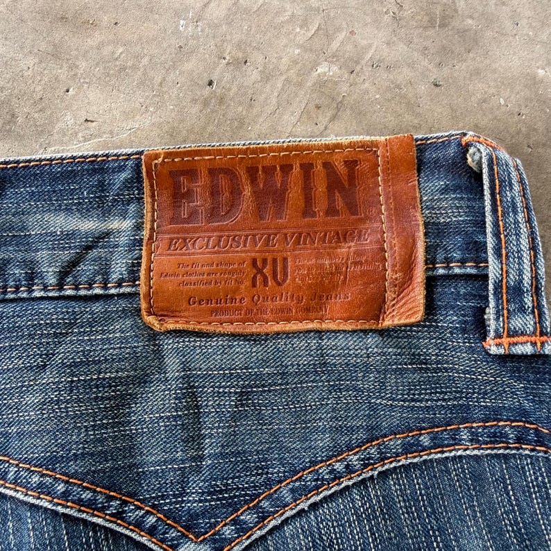 Pu&ograve; includere: Primo piano di jeans di denim blu con cuciture arancioni. Una toppa in pelle marrone con il marchio "EDWIN" e la scritta "EXCLUSIVE VINTAGE" &egrave; cucita sul retro dei jeans. I jeans hanno un classico design a cinque tasche.