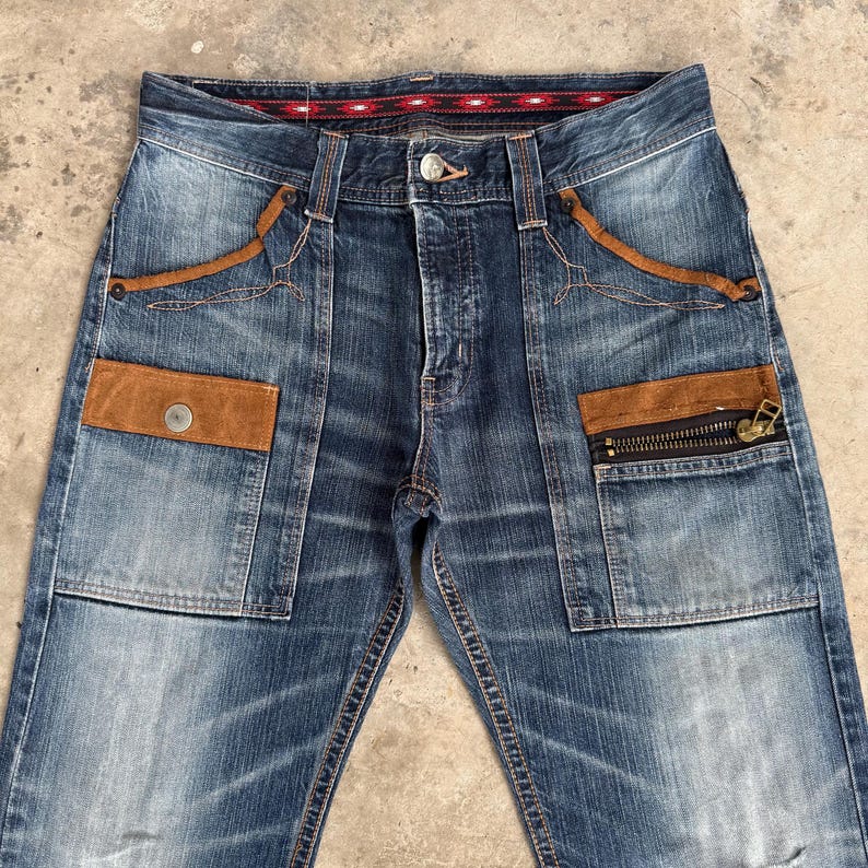 Op de afbeelding: Blauwe spijkerbroek met bruine su&egrave;de-achtige accenten op de zakken en decoratieve stiksels. Voorzien van een knoopsluiting en een zak met ritssluiting. De jeans heeft een vervaagde wassing en een patroon in de tailleband.