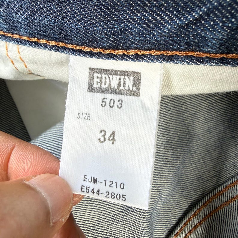 May include: Close-up of a denim jeans label with the brand name "EDWIN" and the number "503". The size is indicated as "34". The label also includes the codes "EJM-1210" and "E544-2805". The jeans are a dark blue wash.