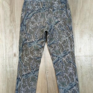 May include: Camouflage cargo trousers with a woodland pattern in shades of green, brown, and grey. The trousers have multiple pockets and drawstrings at the ankles. The trousers are hanging on a wooden hanger.