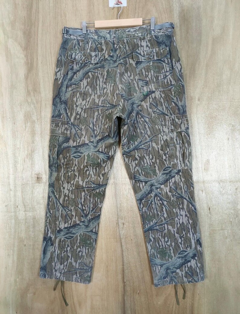 May include: Camouflage cargo trousers with a woodland pattern in shades of green, brown, and grey. The trousers have multiple pockets and drawstrings at the ankles. The trousers are hanging on a wooden hanger.