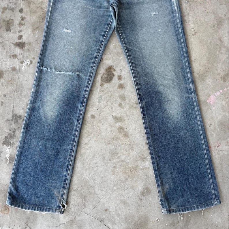 May include: Faded blue denim jeans with a distressed look. The jeans have a straight leg cut, with a worn patch on the left knee and frayed edges at the bottom. The jeans are laid flat on a concrete surface.