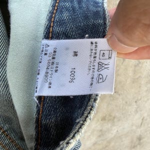 May include: Close-up of a white tag with Japanese text and care symbols, attached to blue denim fabric. The tag indicates the fabric is 100% cotton and provides washing instructions. The tag also includes a phone number.