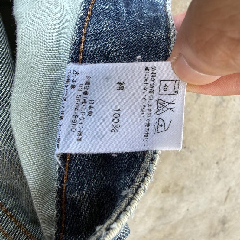 May include: Close-up of a white tag with Japanese text and care symbols, attached to blue denim fabric. The tag indicates the fabric is 100% cotton and provides washing instructions. The tag also includes a phone number.
