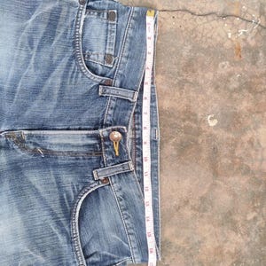 May include: Blue denim shorts with a faded wash, featuring classic five-pocket styling and a button and zipper closure. A measuring tape shows the shorts are approximately 19 inches in length.