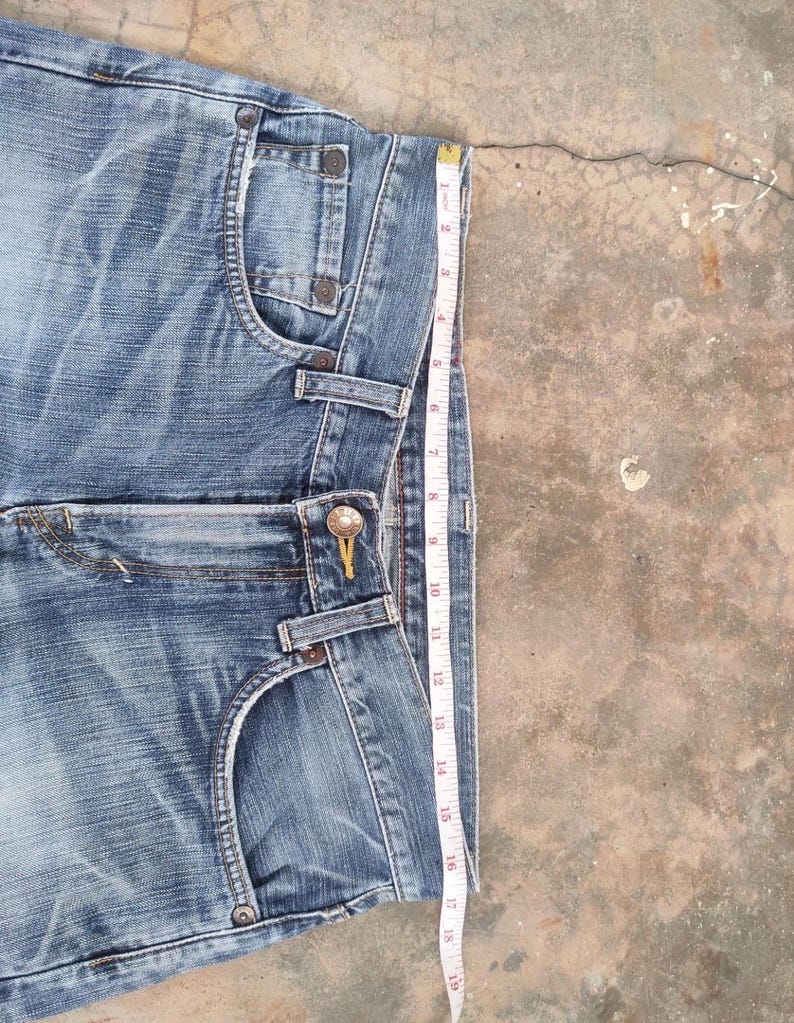 May include: Blue denim shorts with a faded wash, featuring classic five-pocket styling and a button and zipper closure. A measuring tape shows the shorts are approximately 19 inches in length.