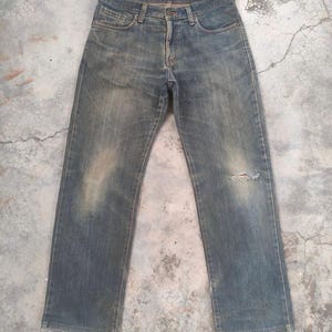 May include: Faded blue denim jeans with a distressed appearance. The jeans have a classic five-pocket design, a button and zipper closure, and a small tear on the right knee. The denim has a worn, vintage look.