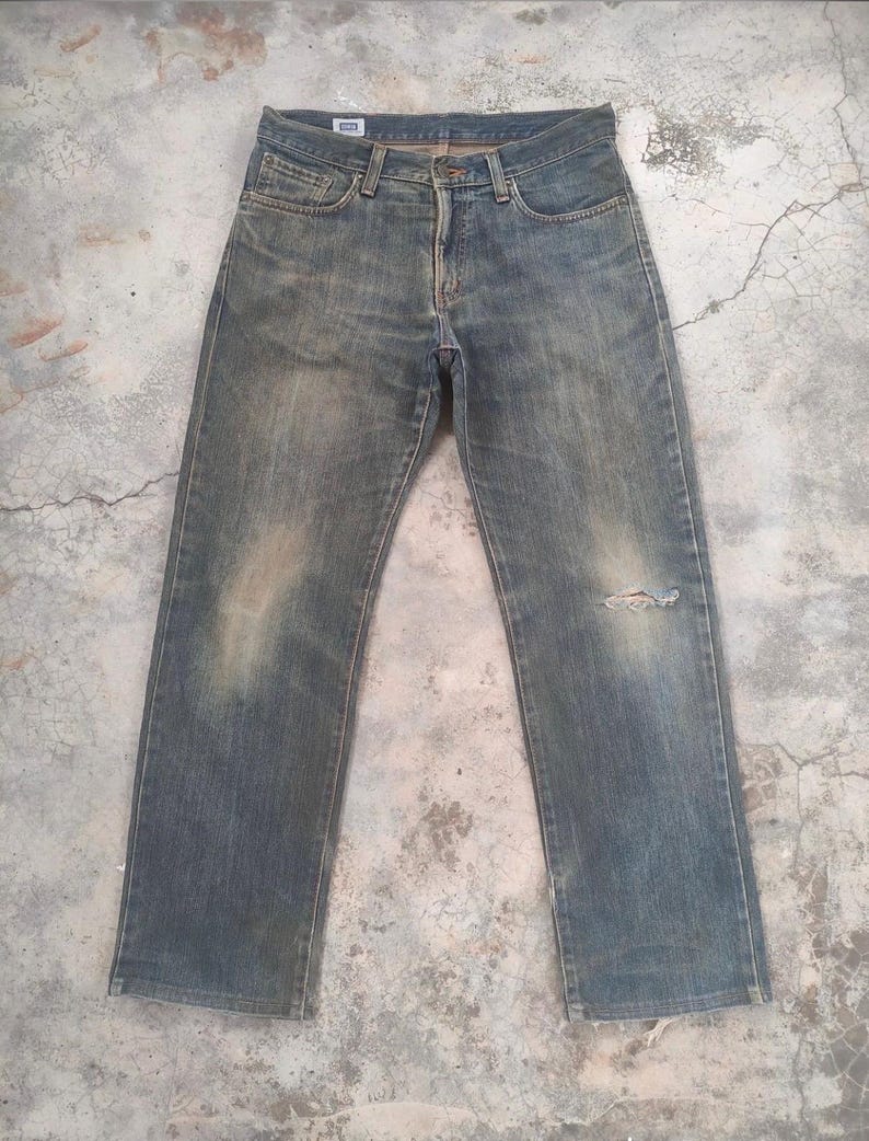 May include: Faded blue denim jeans with a distressed appearance. The jeans have a classic five-pocket design, a button and zipper closure, and a small tear on the right knee. The denim has a worn, vintage look.