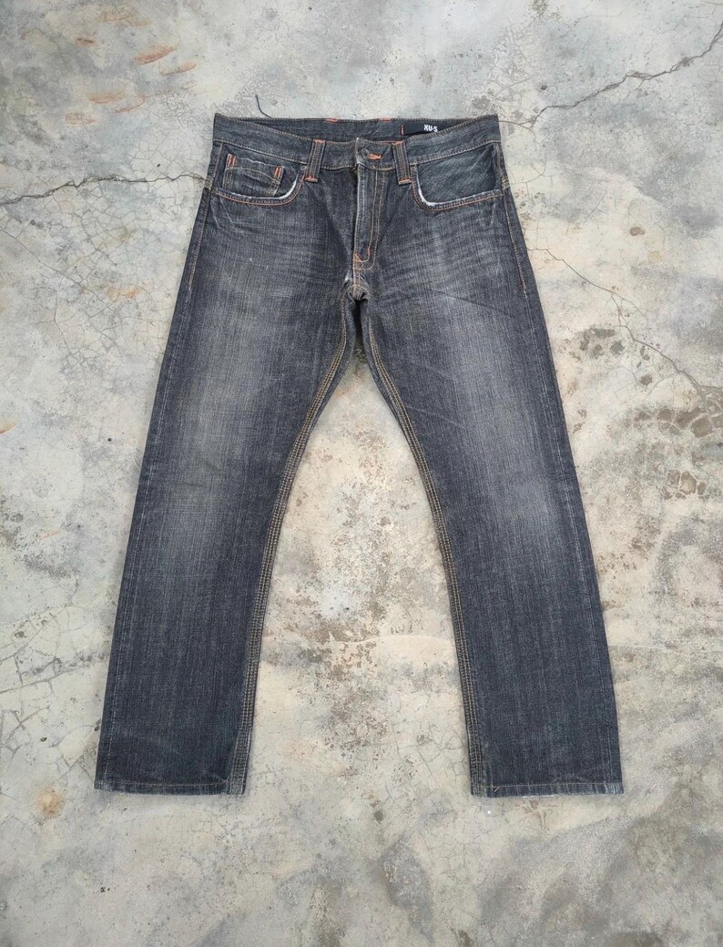 Pu&ograve; includere: Jeans in denim lavato scuro con taglio dritto. I jeans hanno un classico design a cinque tasche, cuciture arancioni e chiusura con bottone e cerniera. Il denim ha un aspetto sbiadito.