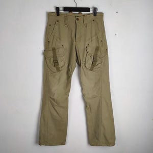 May include: Khaki-colored cargo pants with multiple pockets and a button closure. The pants are hanging on a black hanger against a white background. The pants have two large pockets on the thighs with straps.