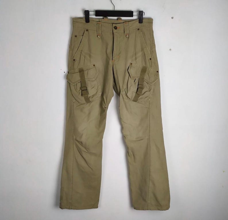 May include: Khaki-colored cargo pants with multiple pockets and a button closure. The pants are hanging on a black hanger against a white background. The pants have two large pockets on the thighs with straps.