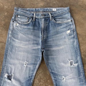 Pu&ograve; includere: Jeans in denim blu chiaro con dettagli strappati, tra cui strappi e sbiaditure. I jeans hanno un classico design a cinque tasche e una chiusura con bottone e cerniera. Il denim appare usurato, suggerendo un'estetica vintage.
