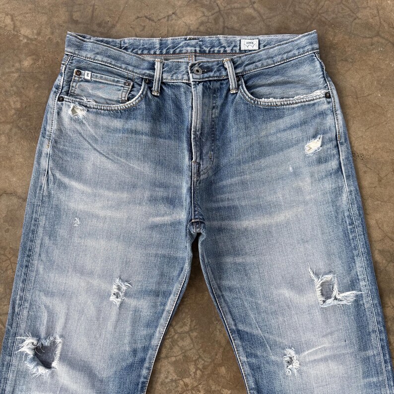 Pu&ograve; includere: Jeans in denim blu chiaro con dettagli strappati, tra cui strappi e sbiaditure. I jeans hanno un classico design a cinque tasche e una chiusura con bottone e cerniera. Il denim appare usurato, suggerendo un'estetica vintage.