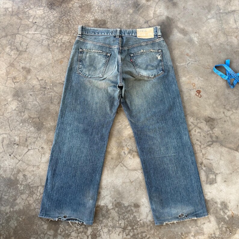 Puede incluir: Vaqueros de mezclilla azul descolorido con un aspecto desgastado. Los vaqueros tienen un dise&ntilde;o cl&aacute;sico de cinco bolsillos, un parche de cuero y una pernera recta. Los bolsillos traseros muestran un dise&ntilde;o desgastado. Los vaqueros est&aacute;n planos sobre una superficie de hormig&oacute;n.