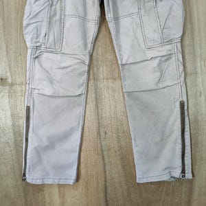 May include: Light gray cargo pants with multiple pockets and zippers on the lower legs. The pants have a slightly worn appearance, with visible stitching and a neutral color palette. The pants are made of a durable material.