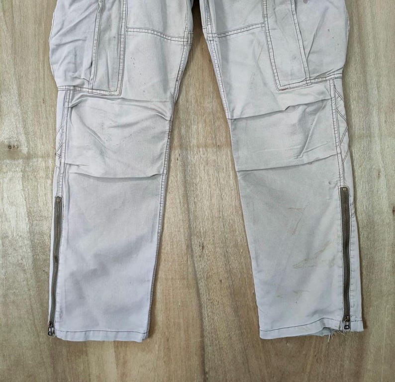 May include: Light gray cargo pants with multiple pockets and zippers on the lower legs. The pants have a slightly worn appearance, with visible stitching and a neutral color palette. The pants are made of a durable material.