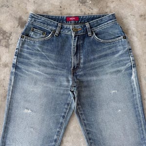 May include: Faded blue denim jeans with a high waist and classic five-pocket design. The jeans have a button and zipper closure, with a small red tag that reads "SOMETHING" above the back pocket. The denim has a worn, distressed look.